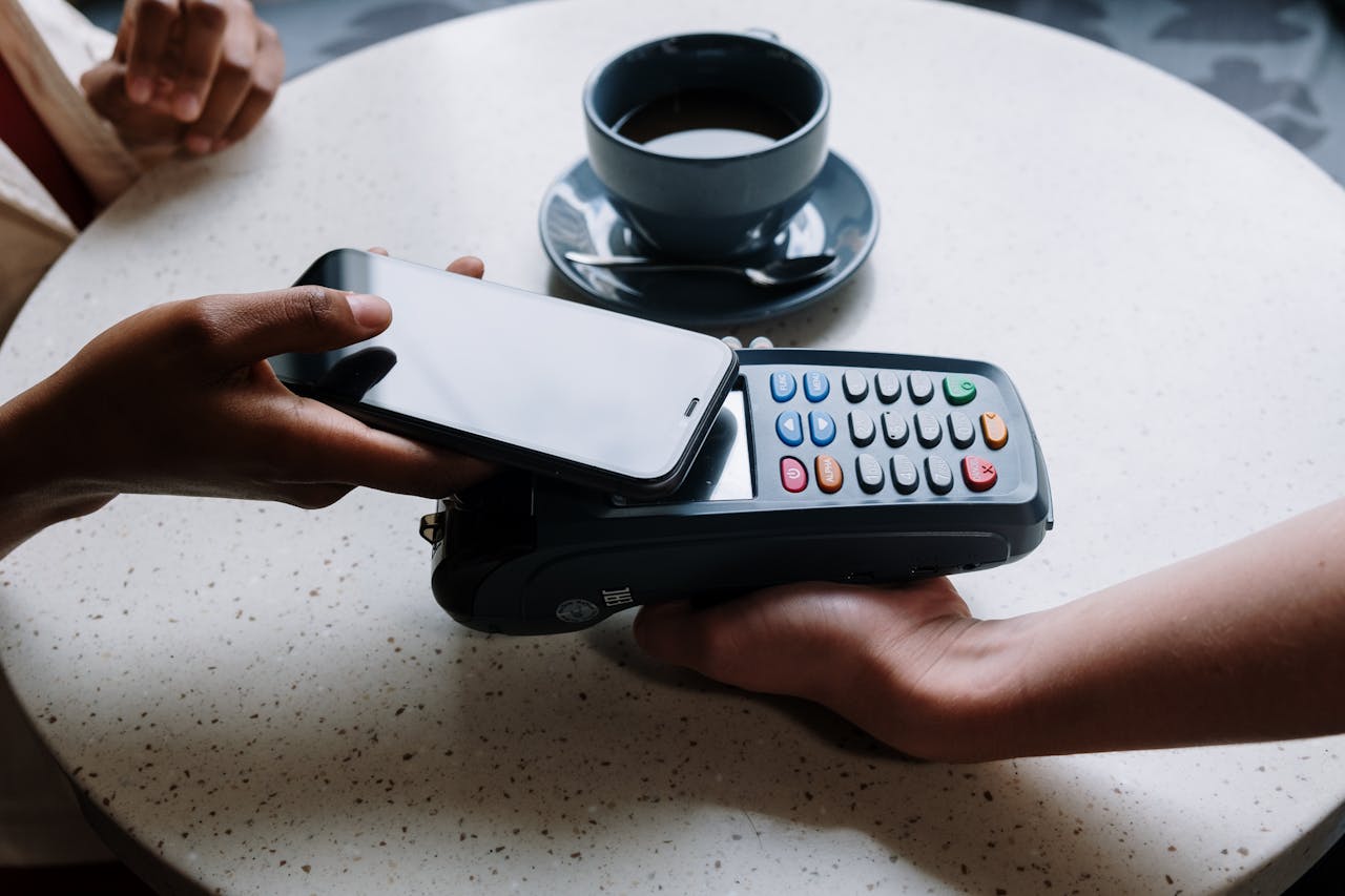 heros-img A person using a smartphone to make a contactless payment with a card reader on a cafe table.